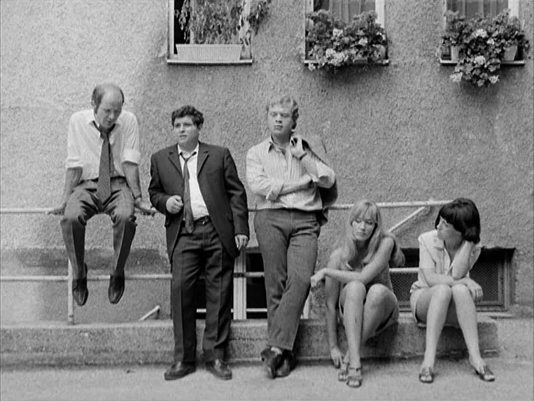 Katzelmacher (1969) movie still: fullbody — Five adults are arranged along a stone ledge against a rough masonry wall. Three window boxes filled…