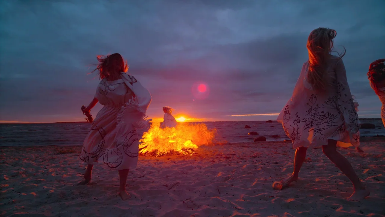 Awaken (2018) movie still: wide — A dark sandy beach at sunset features a large, bright bonfire burning near water. Three women in white patterned…