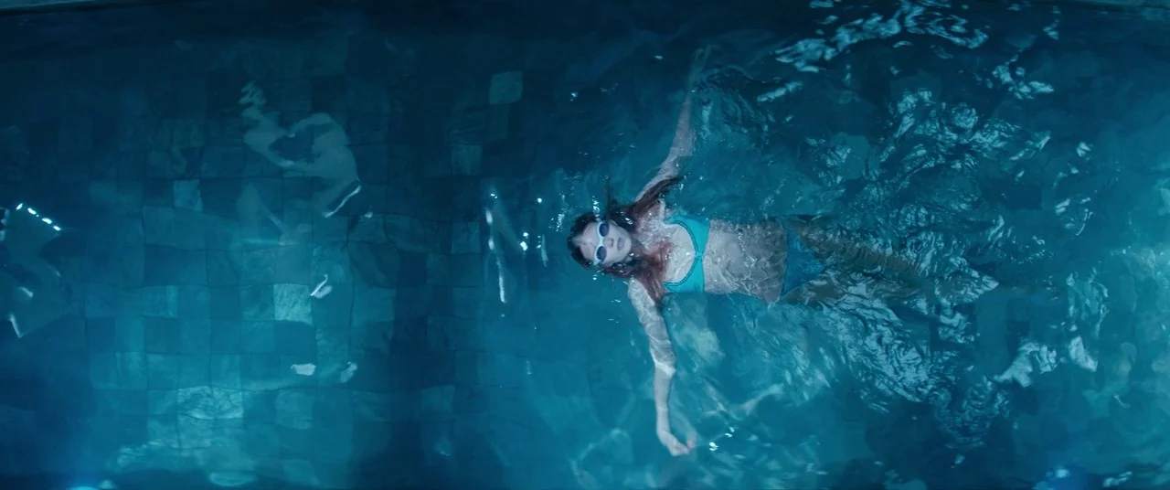 Elizabeth Harvest movie still: wide — A tiled swimming pool filled with dark blue water under subdued lighting. A woman floats motionless on her…