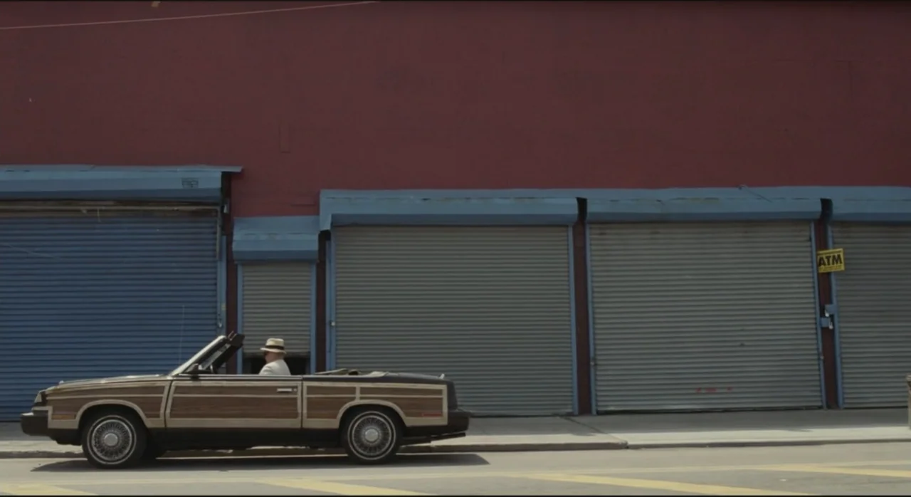 St. Vincent (2014) movie still: wide — A suburban street scene features a red building wall with four closed metal shutters. A wood-paneled…