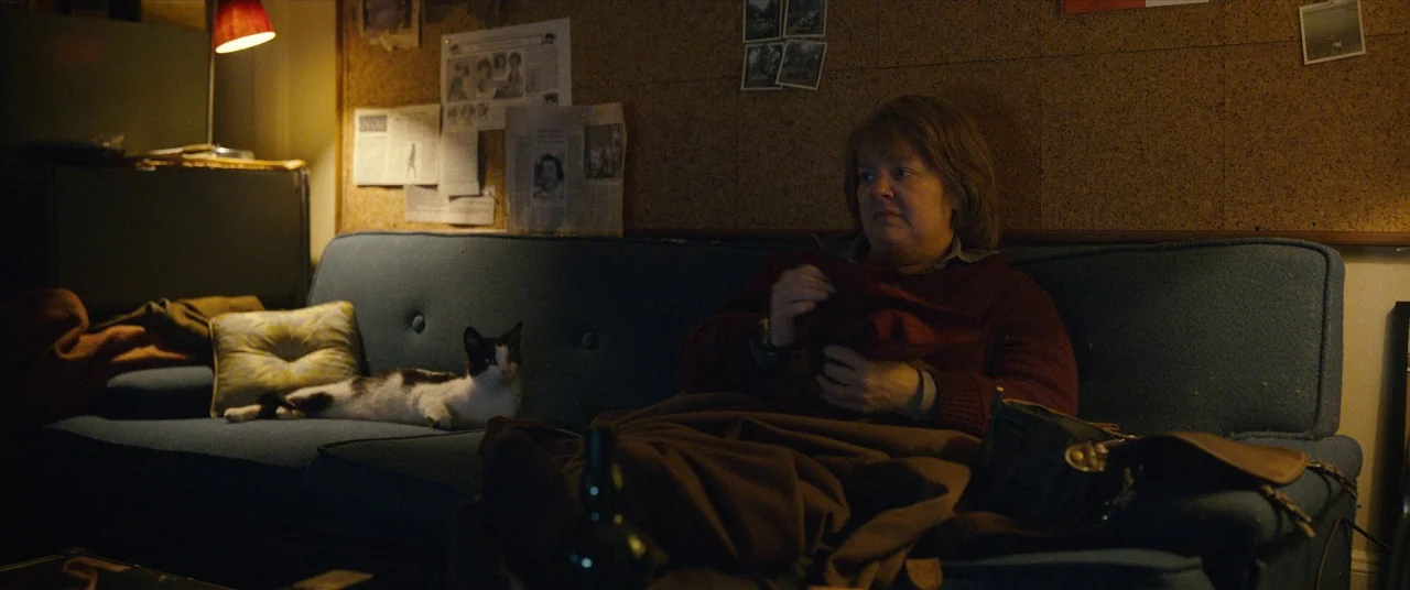 Can You Ever Forgive Me? (2018) movie still: medium — A dimly lit room features a blue sofa against a large wall-mounted cork board. A woman sits…
