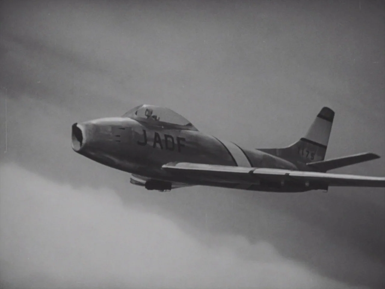 Godzilla Raids Again (1955) movie still: wide — A silver military fighter jet cruises through a cloud-filled sky. The aircraft maintains a steady…