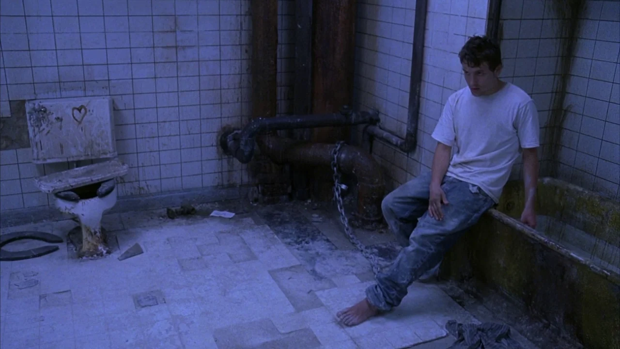Saw movie still: fullbody — A dilapidated, grime-covered bathroom features white square tiles and heavy industrial pipes. A man sits chained to a…