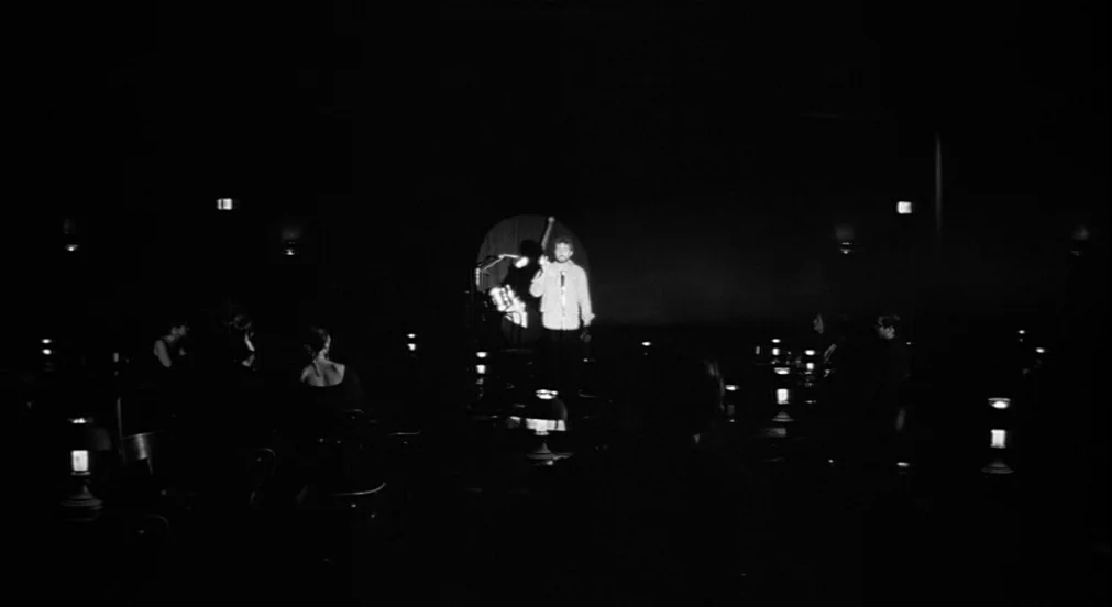 Lenny (1974) movie still: wide — A dim nightclub interior is populated by audience members seated at tables with lamps. A man stands on a central…