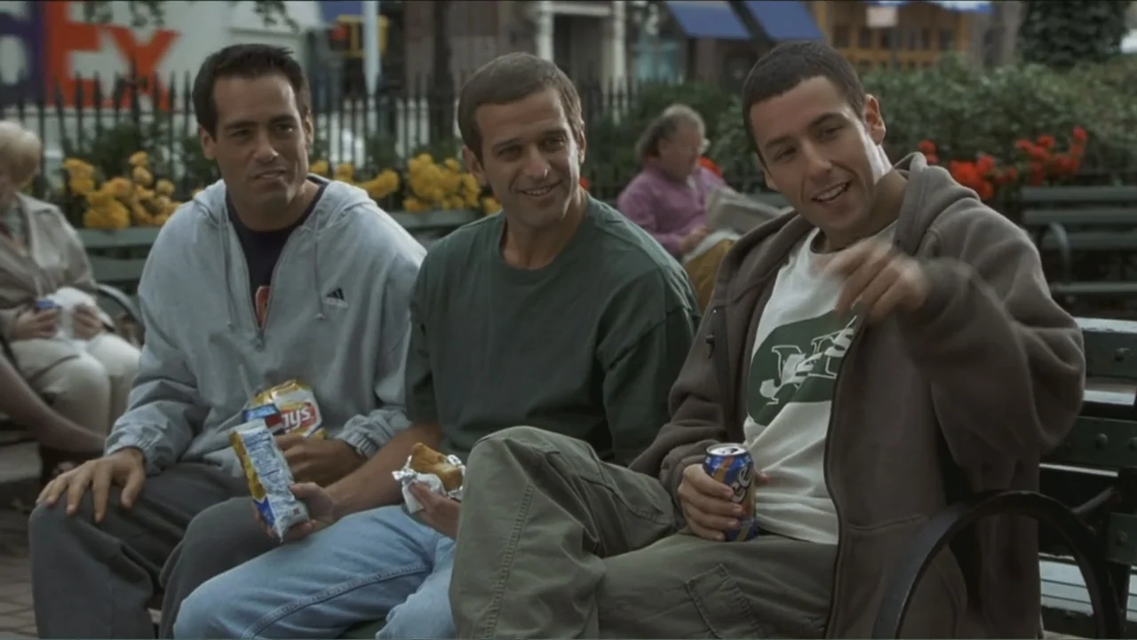 Big Daddy (1999) movie still: medium — Three men sit on a public park bench surrounded by a wrought iron fence. The men talk while holding various…