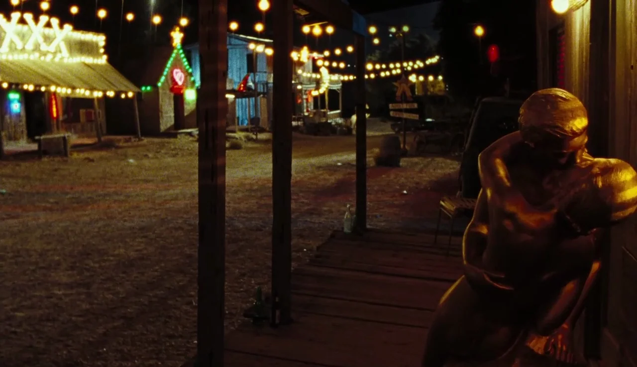 The Devil’s Rejects (2005) movie still: wide — A dusty outdoor set at night features wooden buildings decorated with glowing neon signs. A large…