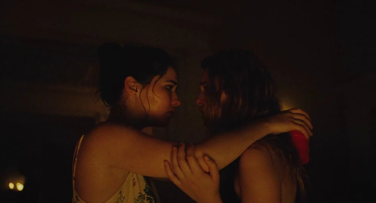 The Giant (2019) movie still: medium — A dark space at night with indistinct surroundings and warm ambient light. Two young women stand face to face…