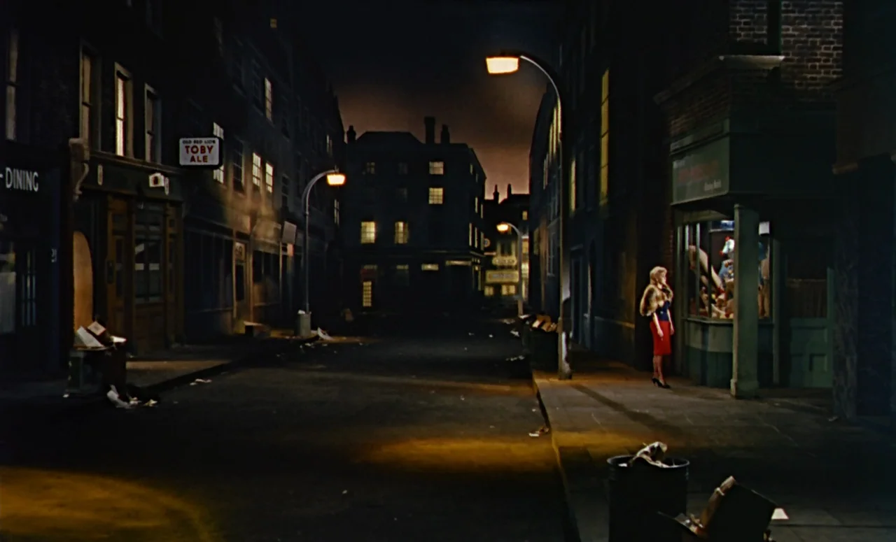 Peeping Tom (1960) movie still: wide — A dark, empty British city street at night lined with multi-story brick buildings. A solitary blonde woman in…