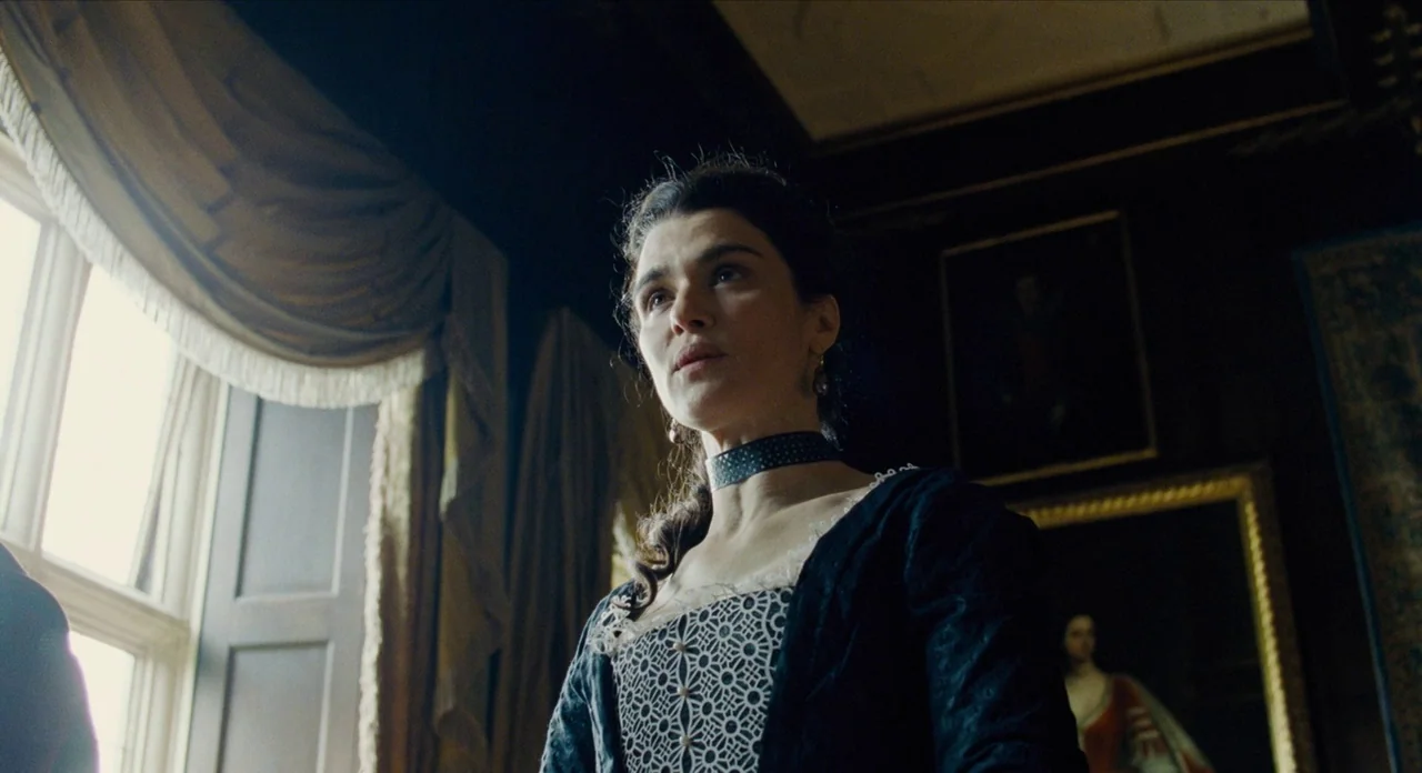 The Favourite (2018) movie still: medium — A dimly lit room with black walls features portraits in heavy gilded gold frames. A woman in a black…