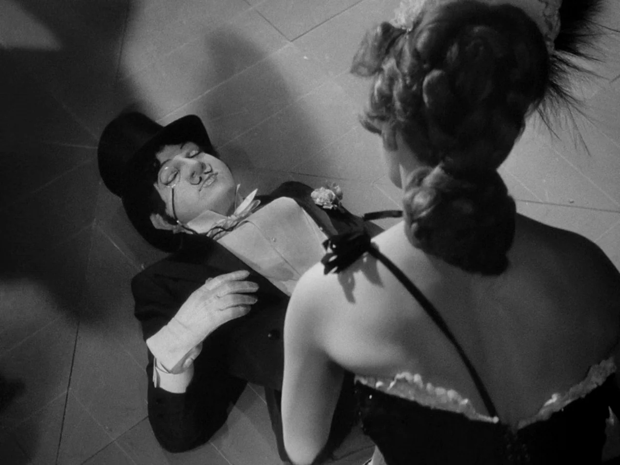 Le Plaisir (1952) movie still: medium — An interior space with a patterned floor surface showing stark light and shadow. A woman in formal dress…