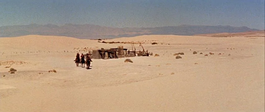 Ride Lonesome (1959) movie still: wide — A vast, open sandy desert expanse features a small wooden structure in the center. Three horseback riders…