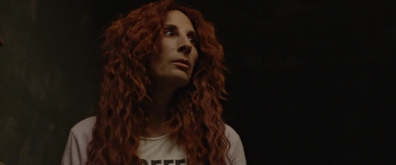 Like or Die (2023) movie still: medium — A dimly lit room with dark walls provides a stark backdrop for the subject. A woman with long red hair…