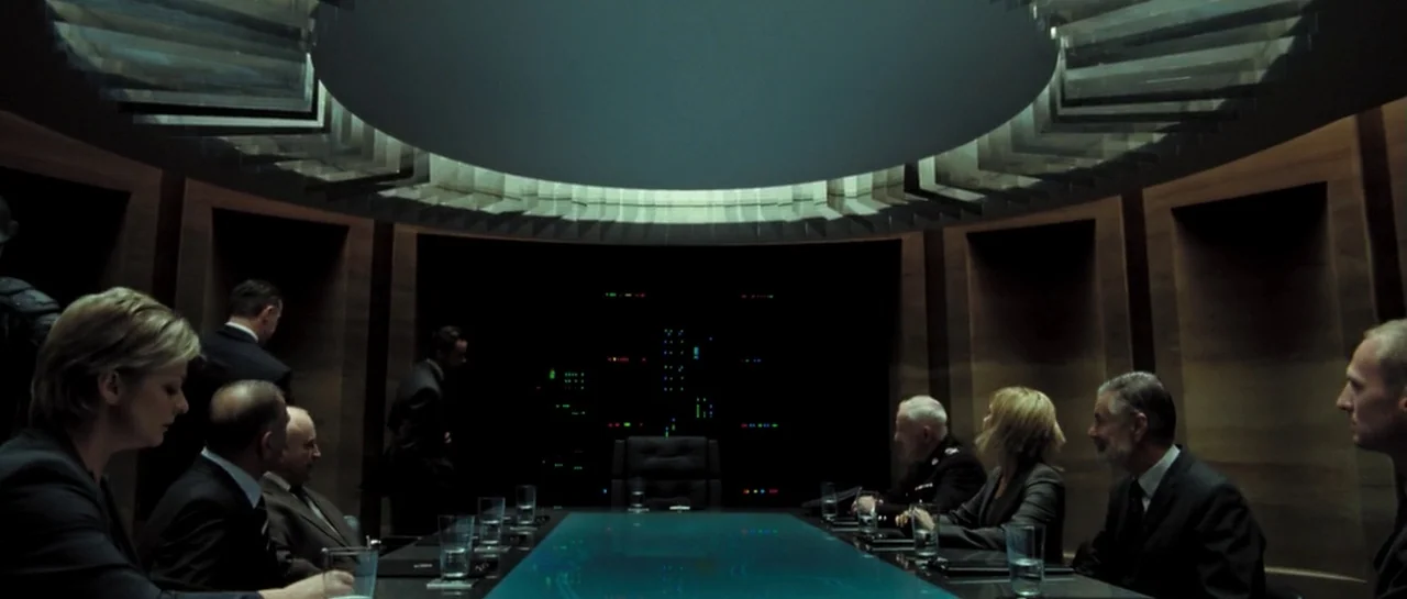 Doomsday (2008) movie still: wide — A dark circular conference room features wood-paneled walls and a large, glowing blue table. Several individuals…