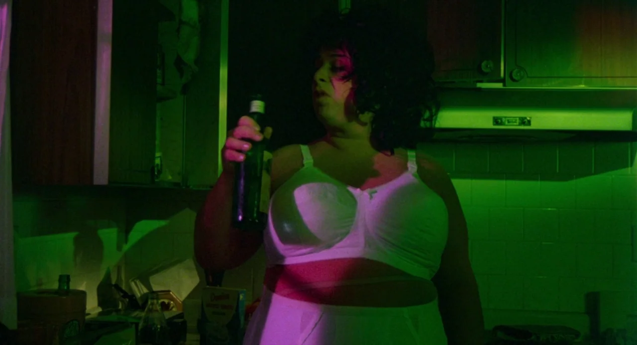 Polyester (1981) movie still: medium — A residential kitchen features dark cabinets, tiled walls, and cluttered countertops at night. A woman in…