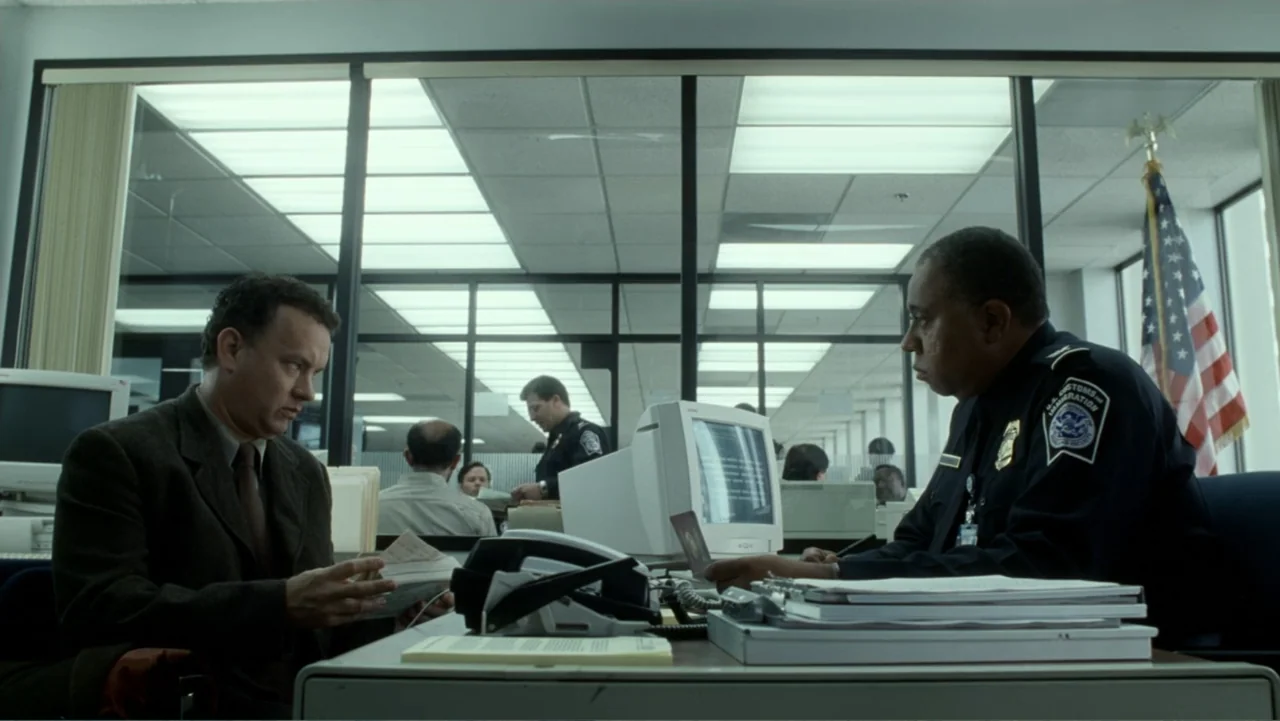 The Terminal (2004) movie still: medium — An airport customs area features glass partitions and rows of computer terminals. A traveler engages with…