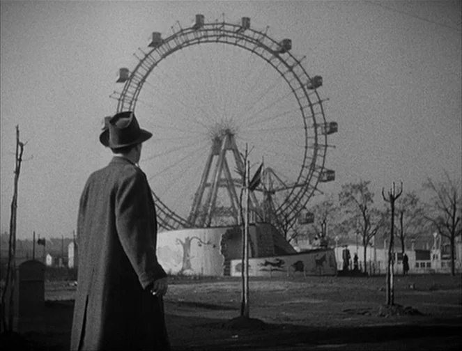 The Third Man (1949) movie still: medium — A man in a long coat stands in an empty, barren park. A massive ferris wheel looms in the background…