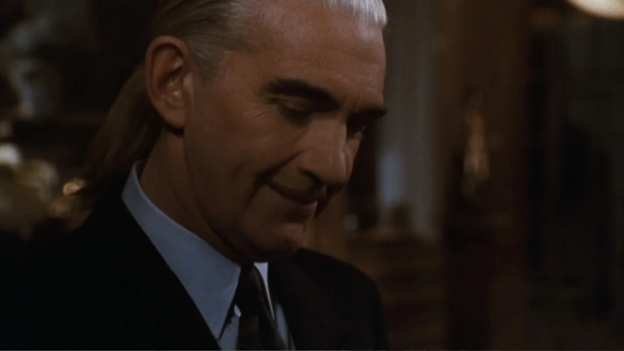 The Bodyguard (1992) movie still: closeup — A dimly lit interior space features an elderly man in a formal dark suit. He keeps his head angled down…