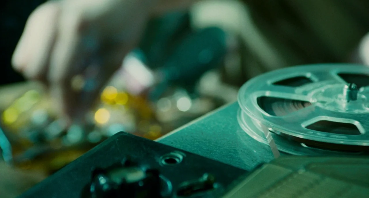 Howl (2010) movie still: closeup — A close-up view of a metal reel-to-reel tape recorder mechanism with dark components. A blurred hand manipulates…