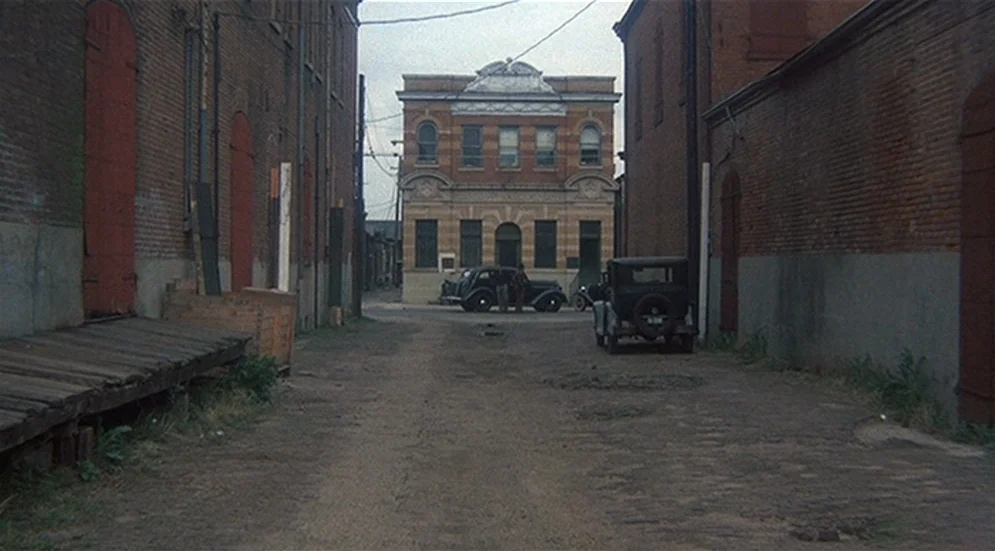 Thieves Like Us (1974) movie still: establishing — An alleyway bordered by high brick warehouse walls leads toward a multi-story masonry building.…