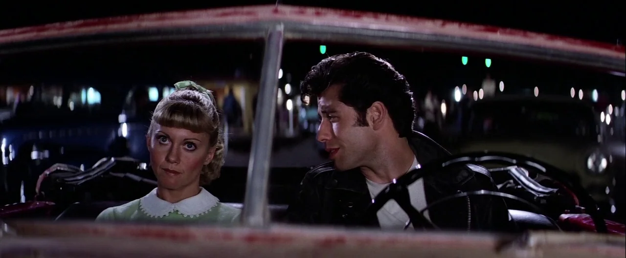 Grease (1978) movie still: medium — A man and woman sit inside an open convertible car at night. The vehicle is parked outdoors with blurry lights…
