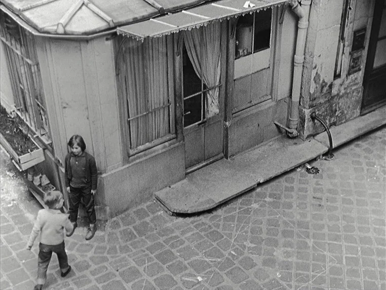 Vivre Sa Vie (1962) movie still: wide — An old corner building sits on a cobblestone street with a faded storefront window. Two young children face…