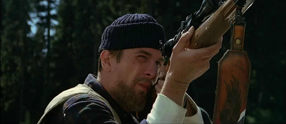 The Deer Hunter (1978) movie still: closeup — A dense forest provides a dark, out-of-focus background for the man in the foreground. The man wearing…