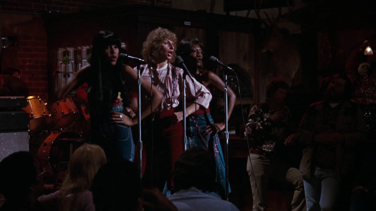 A Star Is Born (1976) movie still: medium — A dimly lit club with a brick wall and a wooden bar in back. Three women stand side by side singing into…
