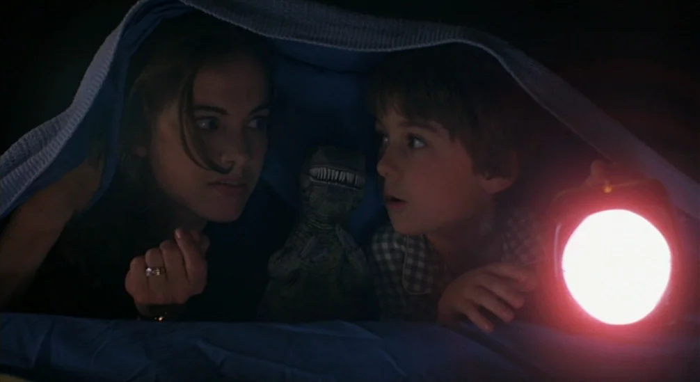 New Nightmare (1994) movie still: medium — A woman and a young boy huddle together under a blue blanket in the dark. The boy holds a glowing…