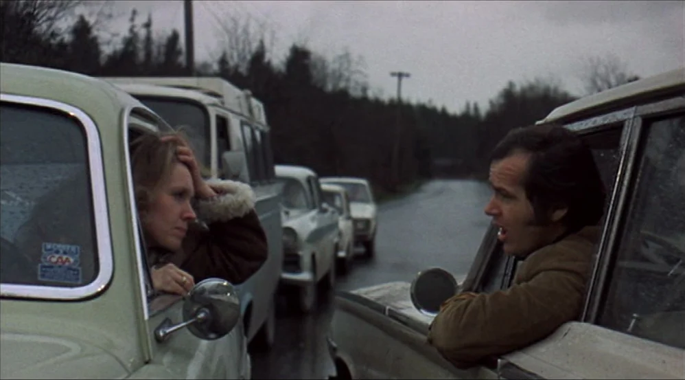 Five Easy Pieces (1970) movie still: medium — An overcast road features multiple vintage vehicles stopped in a queue. A man leans out his…