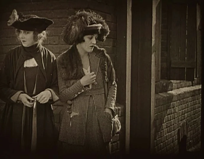 Broken Blossoms (1919) movie still: medium — Two women stand against a weathered brick wall on a bright day. The women remain still while appearing…