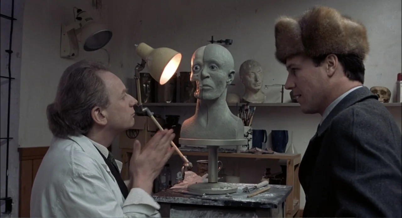 Gorky Park (1983) movie still: medium — A cluttered workshop contains shelves with various busts and tools under dim interior lighting. Two men…