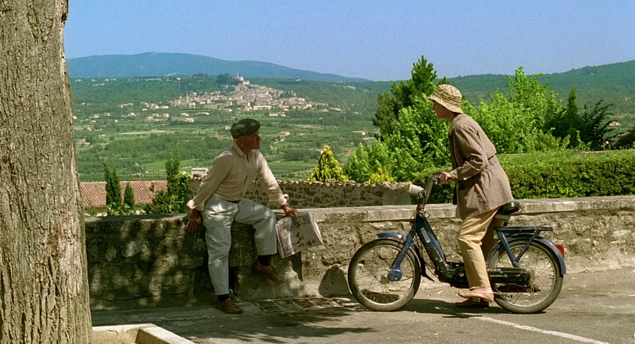 Swimming Pool (2003) movie still: wide — A sunny hillside overlooks a small village nestled in the green valley below. An older man sits on a stone…