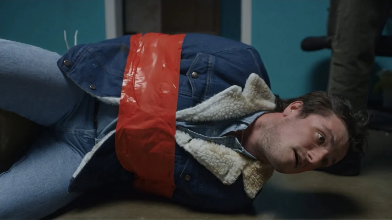 Burn (2019) movie still: medium — A man lies bound on a concrete floor inside a room with teal walls. A man in dark pants stands nearby while the…