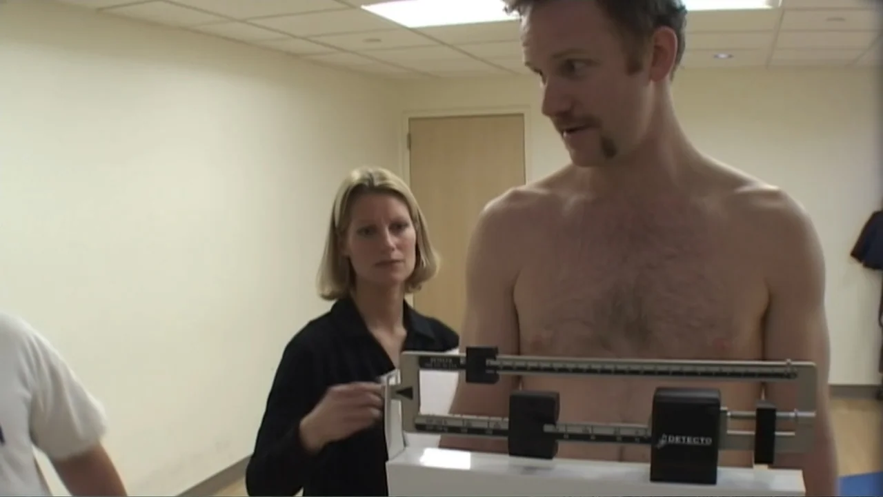 Super Size Me (2004) movie still: medium — A sterile room with off-white walls and a beige door contains a medical scale. A shirtless man stands on…