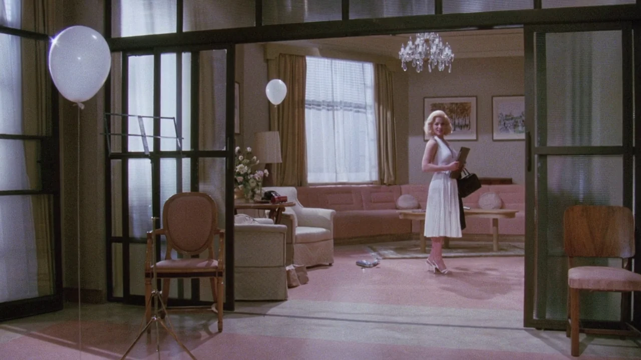 Insignificance (1985) movie still: wide — A living area with pink furniture and glass partition walls. A woman stands in the center of the space…