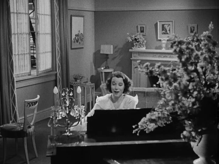A Night at the Opera (1935) movie still: medium — A well-furnished 1930s parlor features a fireplace, framed artwork, and a large bouquet. A woman…