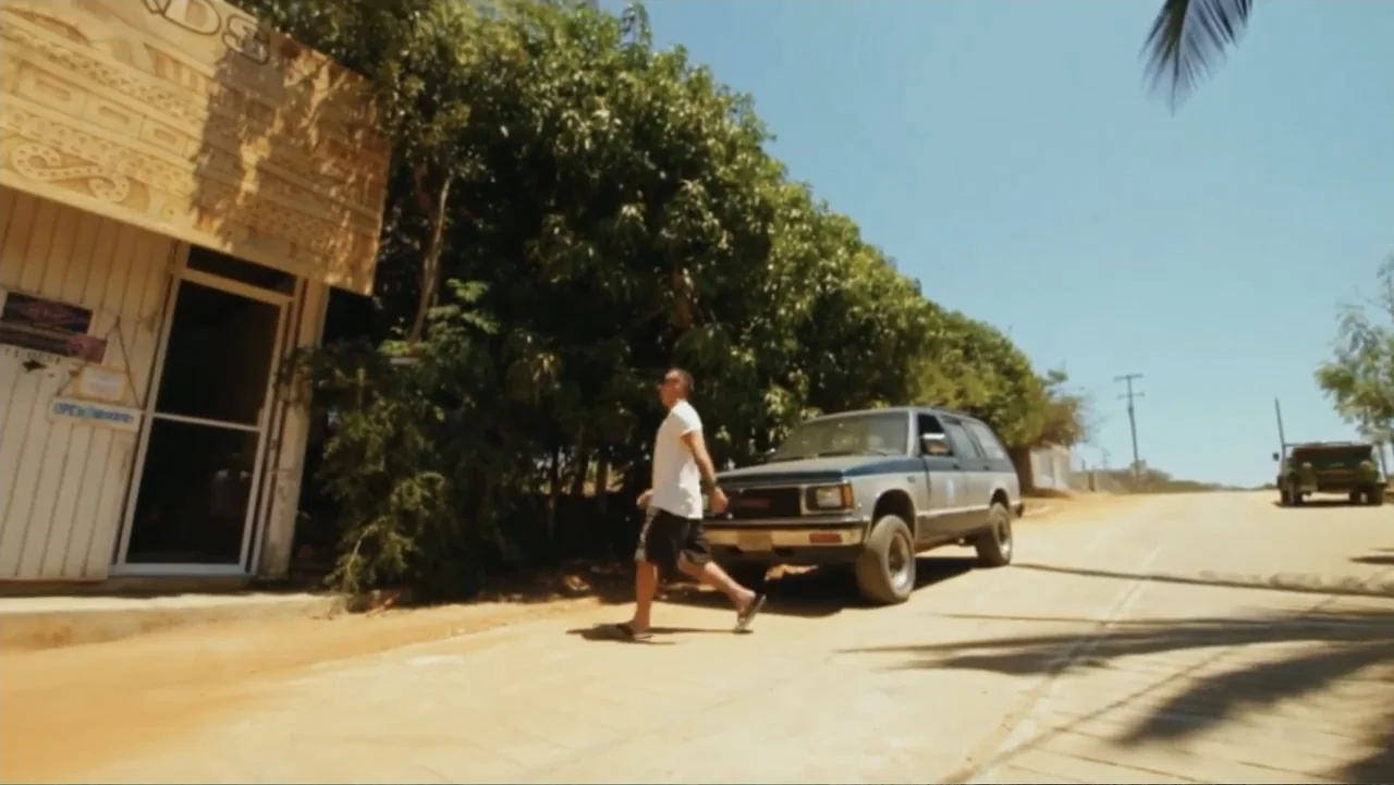 El sueño (2020) movie still: fullbody — A dry, unpaved rural road sits beside a textured stucco building and dense trees. A man walks in profile…