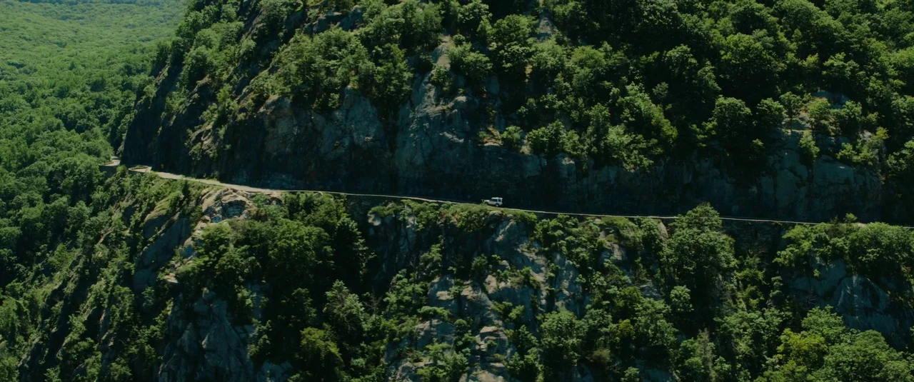 Bodies Bodies Bodies (2022) movie still: wide — A steep mountain slope covered in dense green trees features a thin road. A single white vehicle…
