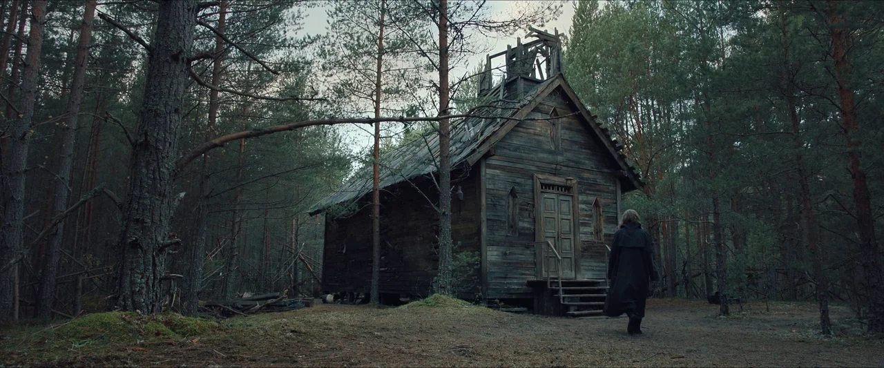 Azrael (2024) movie still: wide — An abandoned, weathered wooden building with a damaged roof stands in a dense pine forest. A lone person wearing a…
