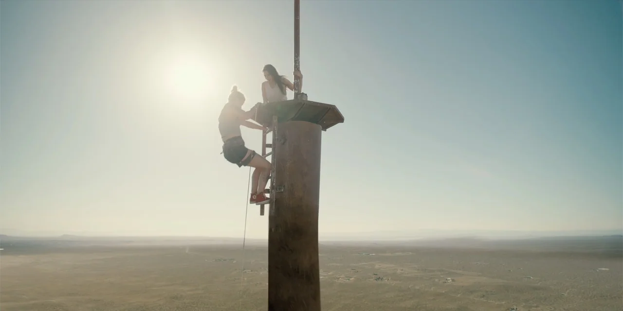 Fall (2022) movie still: wide — A high, metal television transmission tower stands in a vast, arid desert landscape. Two women climb the narrow…
