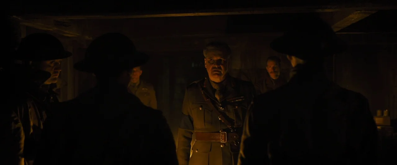 1917 movie still: medium — A dimly lit room with heavy shadows and visible military personnel. An officer in uniform stands centrally as the focus…