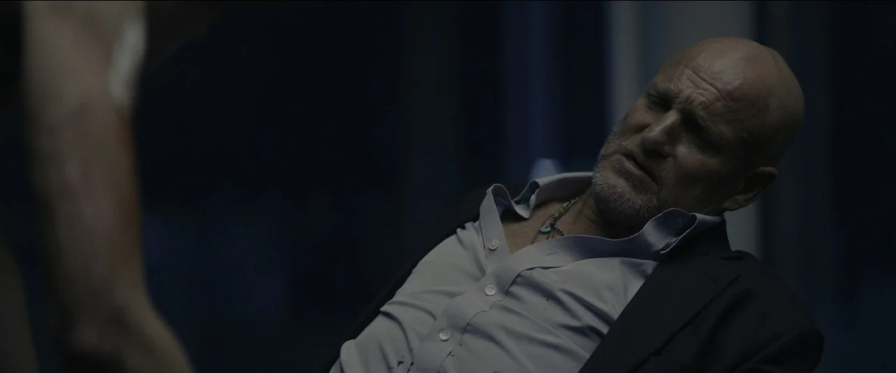 Kate (2021) movie still: medium — A dimly lit room holds a bald man leaning back with his eyes closed. He wears a light-colored dress shirt under a…