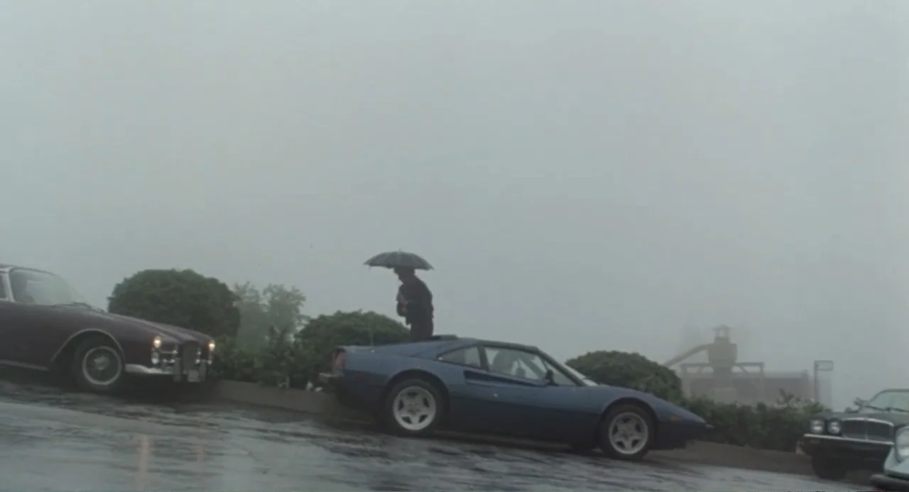 Dangerously Close (1986) movie still: wide — An outdoor parking lot is enveloped in thick grey fog on a rainy day. A man stands beside a blue sports…