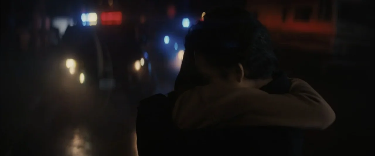 Afraid (2024) movie still: medium — A nighttime city street serves as the backdrop for a close embrace. Two people stand close together with…
