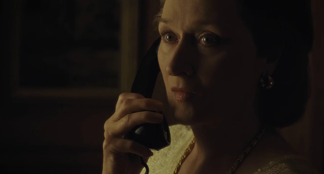 The Post (2017) movie still: closeup — A dimly lit room with dark walls obscured by shadows. A middle-aged woman holds a telephone receiver to her…