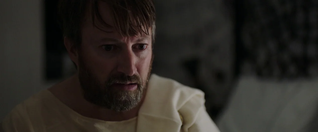 Greed (2019) movie still: closeup — A man in a cream garment looks directly ahead in a darkened room. Soft, directional light illuminates his face…
