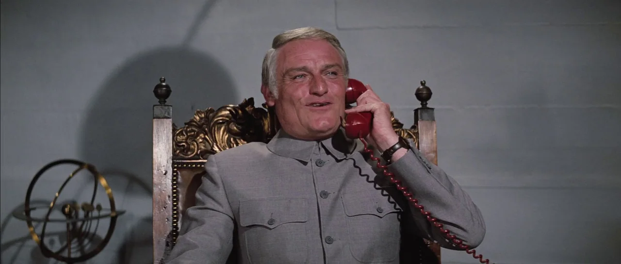 Diamonds Are Forever (1971) movie still: medium — An older man sits in a high-backed ornate wooden chair against a plain wall. He wears a grey tunic…
