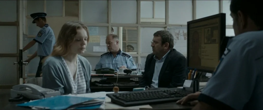 Graduation (2016) movie still: medium — An interior police station office features grey partitions and fluorescent lighting with office furniture. A…