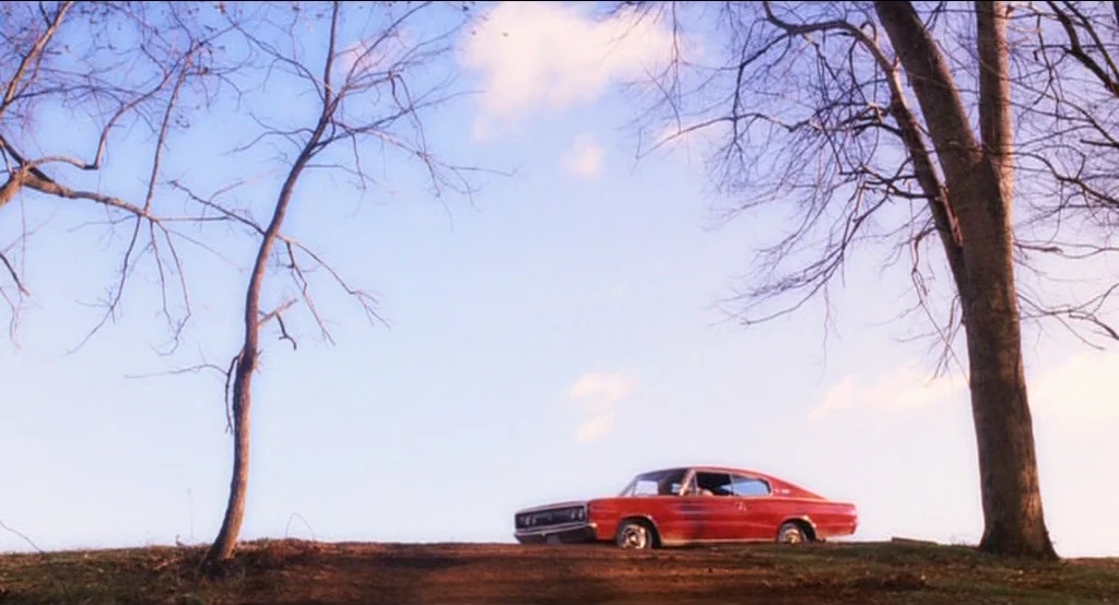 Big Fish (2003) movie still: wide — A red vintage sedan is parked on a grassy dirt mound against a blue sky. Two leafless trees frame the vehicle on…