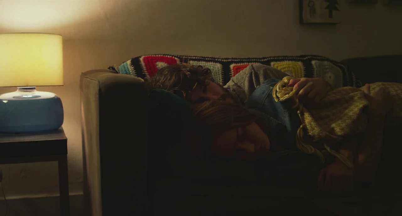 Short Term 12 (2013) movie still: medium — A dark, dimly lit room contains a sofa where two people are sleeping soundly. A lit table lamp stands to…